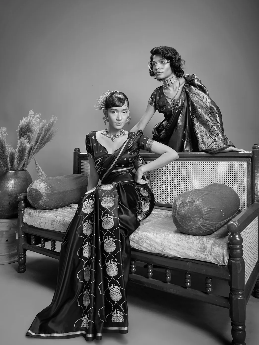 Black-and-white fashion photograph of two models wearing sarees beside a cane sofa.